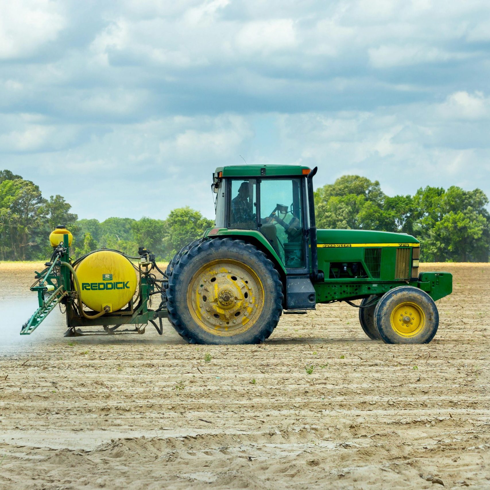 Green tractor with sprayer working in an open farm field under a cloudy sky.